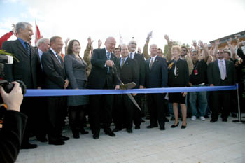 Photo of a man cutting a wide ribbon with a pair of oversized scissors. He is flanked by several men and women in business suits with a crowd of college students behind him and the Capitol Building visible in the background. 