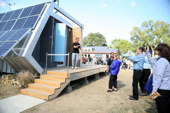 Photo of a young man standing on a deck outside of a futuristic-looking house talking to a group of five people standing at ground level.