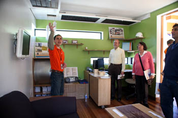 Photo of a young man in an orange T-shirt pointing to a feature inside the office of a home, with three people looking on.