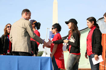 Photo five young women in red T-shirts walking in procession. One is about to shake the outstretched of a man in a business suit. The Washington Monument is visible in the background.