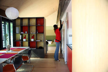 Photo of the interior of a house. A young woman is to one side reaching up and making an adjustment to a shelf. Large blocks and bands of color are part of the décor.  