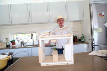 Photo of a man in a cowboy hat standing behind a model of a house that's on a kitchen counter.