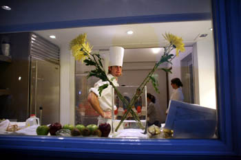 Photo of a young man in a chef's toque standing in a kitchen. He is visible through the window of the house.