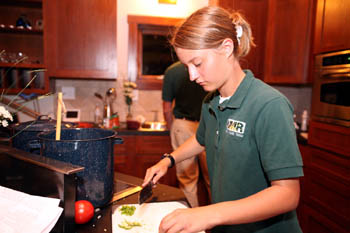 Photo of a young woman in a green polo shirt standing at a stove and chopping vegetables. 