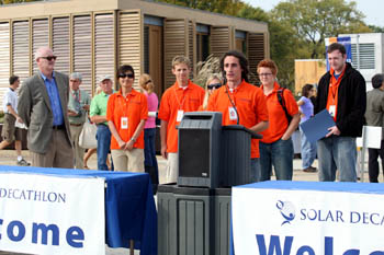 Photo of six people standing behind a table with a podium. Five of them, including the speaker, are wearing orange polo shirts.