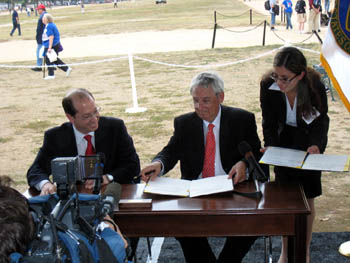 Photo of two men in black business suits and red ties sitting down and signing an official-looking document. A woman stands to their right.