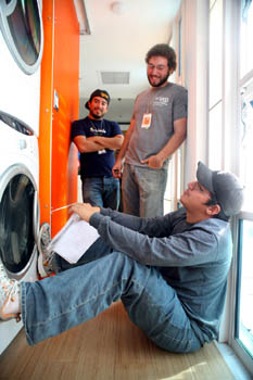 Photo of a young man sitting down in a hallway with his feet up on a washing machine and two young men standing and looking down at him. All three are smiling. 