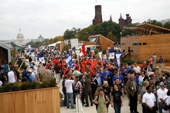 Photo of a large group of people, several in team uniforms and some carrying team banners, walking down the pathway between the solar houses