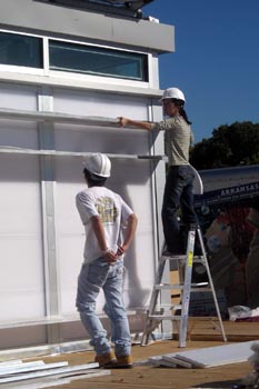 Photo of a woman in a hard hat on a ladder and a man below her, both working together to remove a longitudinal plastic panel from the side of the solar home.