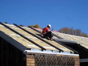 Photo of a man with a hard hat kneeling on the roof of a solar home using a wrench on a solar photovoltaic panel.