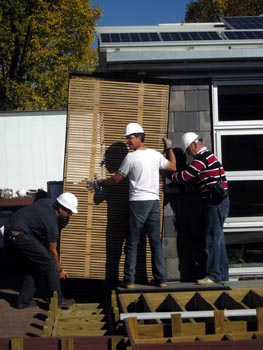 Photo of two men with hard hats lifting a lattice-work wooden panel from the side of a solar home.