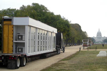 Photo of a section of a solar house sitting on a trailer, lined up behind four other trucks with house sections on their trailers. The U.S. Capitol building is in the distance