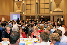 Photo of people sitting at round tables in a large ballroom, listening to a presentation.
