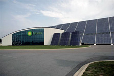 Photo of a building with two sections. One section is white, has a the BP Solar sign and logo, and has a roof curving from the ground to halfway up the other section, which is a slanting wall covered with solar panels. Three more large solar arrays are in front of the slanted wall.