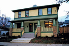 Photo of a yellow older-style duplex house with a front porch across the front, a small yard, and green trim.