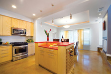 Photo of a bright kitchen with light wood cabinets, red countertops, stainless steel appliances, and two glass doors letting in light.