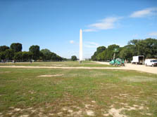 Photo of the National Mall. The Washington Monument can be seen at the end of a long grassy area ringed by walkways and trees. A few work trucks and vans, as well as a snorkel lift are parked on the walkway to the right.