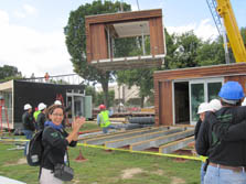 Photo of a woman clapping while a crane lifts part of a house in the air. The module hovers over the main part of the house. Small groups of people look on.
