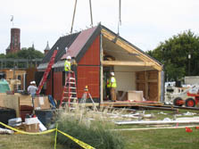Photo of a house under construction. Workers are standing inside a portion of the house that is without a wall. Another is on a ladder inspecting the roof.