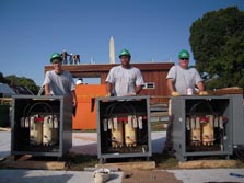 Photo of three men wearing green hard hats and standing behind open metal boxes that come up to their waists. Instrumentation is visible inside the boxes. In the background, the peak of the National Monument juts above a house under construction.