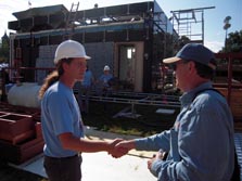 Photo of two men shaking hands. In the background is North House, a glass house under construction.