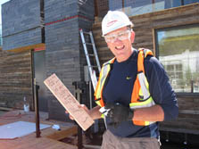 Photo of a man holding a one-foot section of a wood 2 x 4 with writing on it. He is smiling and holding a pen. He is standing on a construction site.
