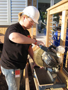 Photo of a woman wearing a hard hat. She is using a power saw to cut pieces of wood.