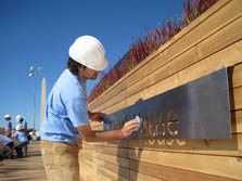 Photo of a man wearing a hard hat and wiping off a sign attached to a wooden retaining wall. The Washington Monument is in the background.