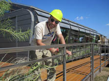 Photo of a man wearing a hard hat and filing a metal railing. He is standing on a deck.