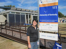 Photo of a young woman standing next to a sign in front of a house. There is a long ramp to the house behind her.