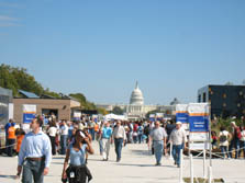 Photo of a crowd of people walking and looking at houses on either side of a wide walkway. The U.S. Capitol is in the background.