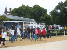 Photo of people outdoors, many with umbrellas or hats, and a modern-looking house in the background with a roof that appears to float.