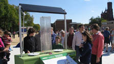 Photo of a small group of adults and children gathered around an exhibit with a plexiglass-encased water fountain underneath a solar panel.