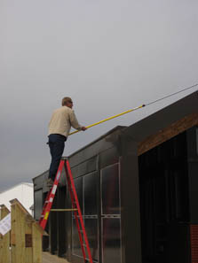 Photo of a man standing on a ladder next to a sloped roof. He is using a long-handled tool to clean off solar panels.