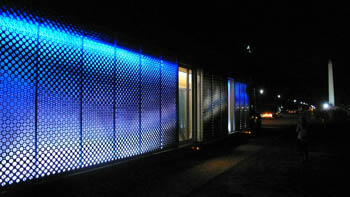 Photo of a diesel carrying a rectangular building on its flatbed trailer. The side of the building is illuminated in blue light. The Washington Monument is visible in the distance.