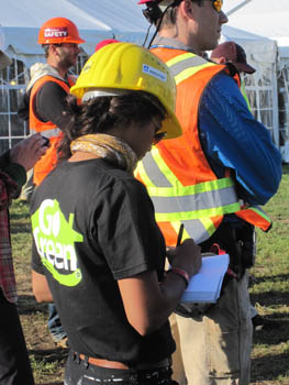 Photo of a young woman in a hard hat taking notes in a small notebook while standing. Behind her are several men wearing hardhats and safety vests.