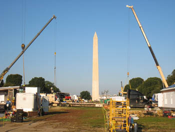 Photo of the National Mall during construction of the solar village. Construction equipment and materials can be seen in the foreground. On the sides, two extended cranes frame the Washington Monument, which can be seen in the distant background.