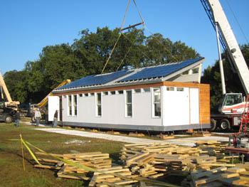 Photo of a small white rectangular house. A crane behind the house is lowering the roof, which is covered in photovoltaic panels, into place. In the foreground are stacks of wooden braces.