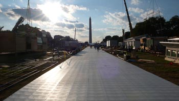 Photo of a white walkway lined on both sides by houses under construction. At the end of the walkway is a white tent, and behind that is the Washington Monument.