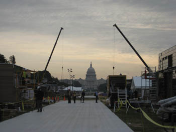 Photo of solar village construction on the National Mall. Team houses line a central walkway. In the background, extended cranes frame the U.S. Capitol.