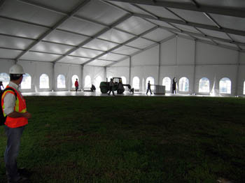 Photo of six people wearing hardhats inside a large white enclosed tent. They are installing white flooring, which extends only part way over the grass inside. A man wearing a hardhat and safety vest looks on in the foreground.