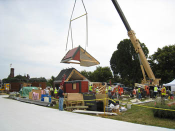 Photo of a large crane lowering a roof onto an unfinished house. Numerous people wearing hardhats and safety vests watch the process.