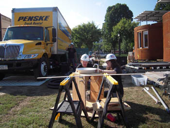 Photo of two women kneeling around a wooden cabinet assembly that has a white porcelain sink bowl on top. To the right, the corrugated steel cladding on the home appears oxidized. To the left of the women is a large yellow rental truck.