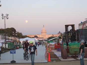 Photo of the moon rising next to the capitol building. In the foreground, people stroll and bike down Decathlete Way, which is surrounded by construction.
