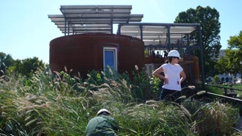 Photo of a young woman wearing a hard hat and gazing over her shoulder. Another person works on the tall grasses in front of a cylindrical house.