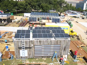 Aerial photo of the roof of a house, with solar panels angled toward the sun. Three men in hardhats and safety gear are installing a panel.