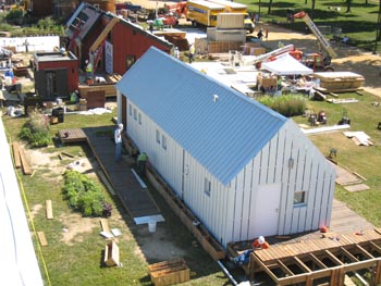 Aerial photo of a house that resembles a small barn. Surrounding the house are construction materials and other houses under construction.
