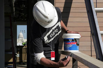 Photo of a person wearing a Team Boston t-shirt and a hard hat applying spackle to a wood railing. An open door behind shows through to a window and, behind that, the U.S. Capitol.