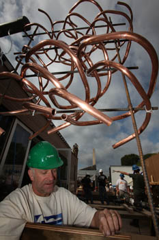 Photo of a man wearing a green hard hat installing copper pipes. Above his head, a metal structure holds numerous winding copper pipes.