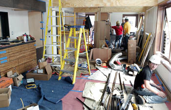 Photo of several people working inside an unfinished team house. Construction materials , ladders, blankets, and boxes are strewn about inside.